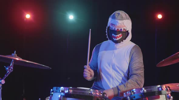 A Male Musician Disguised As a Shark Plays a Drum Kit in a Dark Studio with Bright Colored Lights