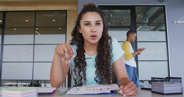 Biracial creative businesswoman at desk, reading, making notes and talking during video call