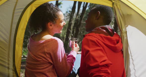 Smiling diverse couple sitting in tent and drinking tea in countryside