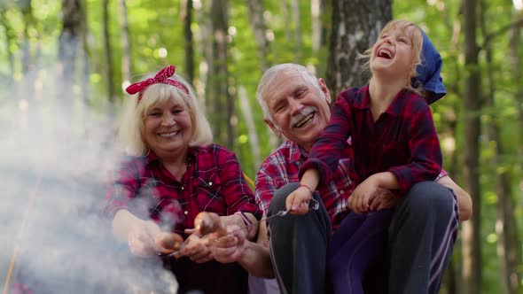 Senior Man Woman with Granddaughter Resting at Camping in Wood Cooking Frying Sausages Over Campfire