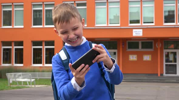 A Young Boy Plays a Game on a Smartphone with a Smile in Front of an Elementary School