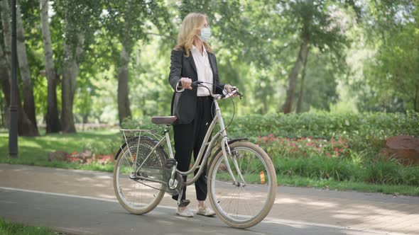 Wide Shot of Confident Caucasian Middle Aged Woman in Suit and Coronavirus Face Mask Sitting on Bike