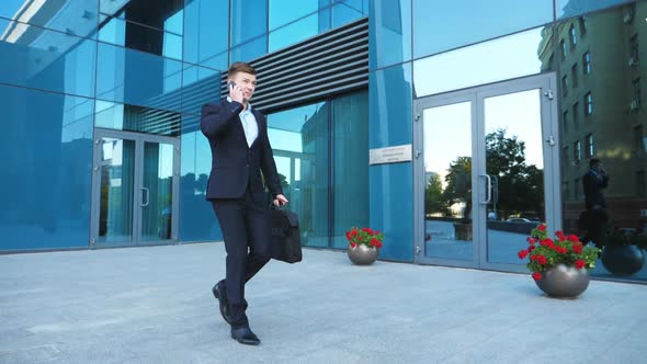 Young Businessman in Suit Walking Along City Street and Talking on Phone Near Modern Building
