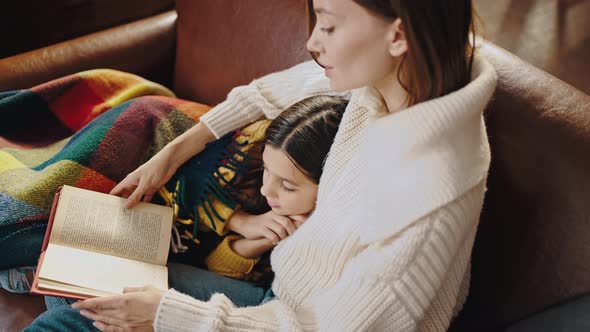 Young Mother Reading Book to Preteen Daughter Covered By Checkered Plaid While Sitting on Sofa at