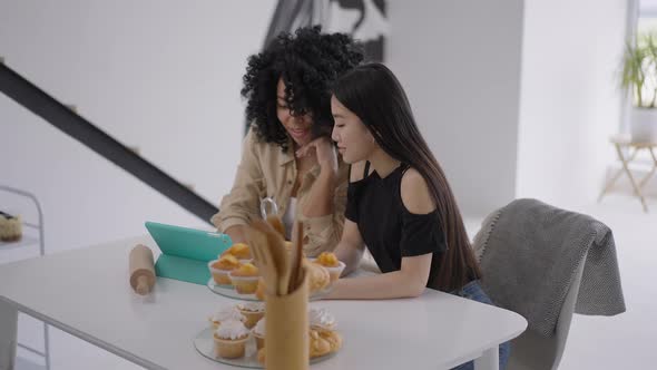 Laughing Beautiful Young Women Talking Surfing Internet on Tablet Sitting at Table with Freshly