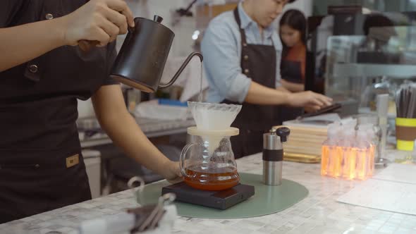 A female café operator wearing an apron pours hot water over roasted coffee grounds to prepare.