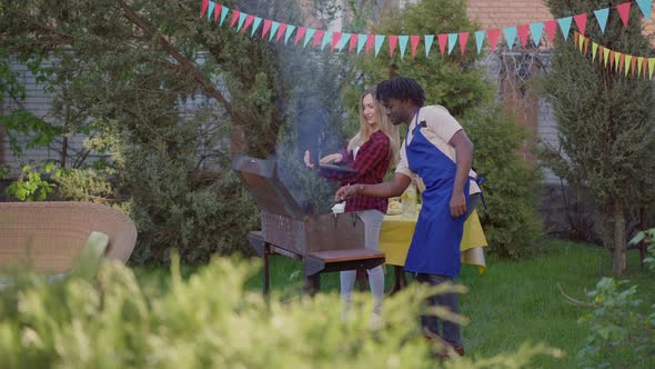 Wide Shot Side View of Young African American Man Preparing Barbecue As Caucasian Woman Showing