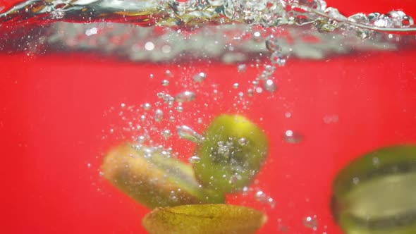 Slices of Fresh Cut Kiwi Fall Down in Clear Water with Bubbles on Red Background