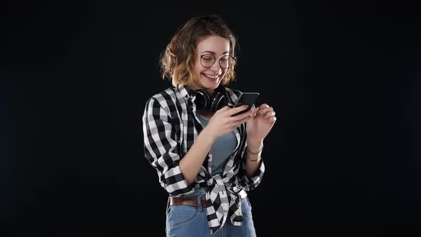 Cheerful Woman Wearing Casual Clothing Plaid Shirt and Jeans Holding Smartphone and Scrolling the