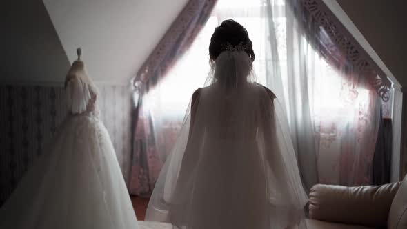 Unrecognizable Bride in White Veil Walks to Window Past Wedding Dress