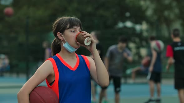 A Teenage Girl in a Lowered Medical Mask Stands on a Street Sports Basketball Court with a Ball