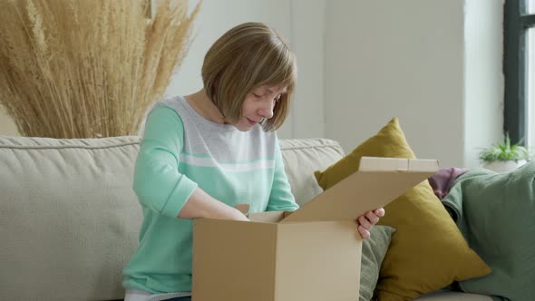 Middle Aged Woman Unpacking Cardboard Box Sitting on Sofa at Home