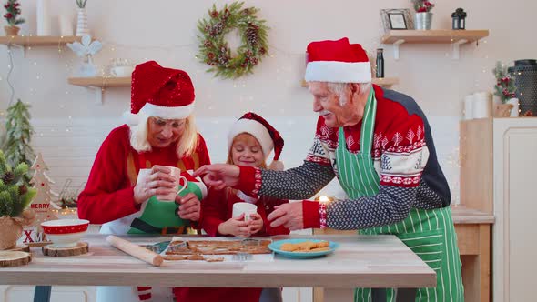 Senior Man with Cups Hot Chocolate Walking Into Christmas Home Kitchen to Grandmother and Grandchild