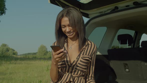 Positive Pretty African Female Tourist in Car Trunk Networking on Cellphone