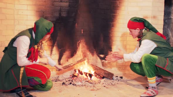 Elves in green fairytale costumes are sitting by a large fireplace with fire.