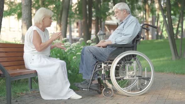 Wide Shot of Senior Caucasian Woman in White Dress Sitting on Bench in Summer Park and Talking To