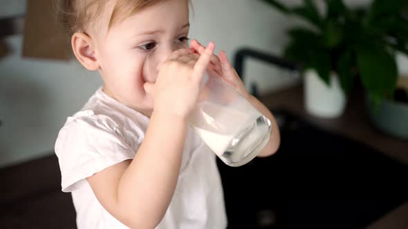 Happy Baby Girl Sitting at the Table in the Kitchen and Drinking Milk