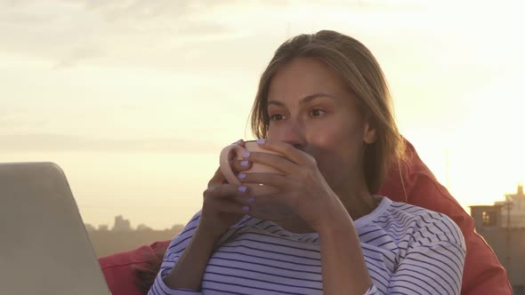 A Resting Girl Drinks Warm Tea and Looks at the Laptop Sitting on the Roof