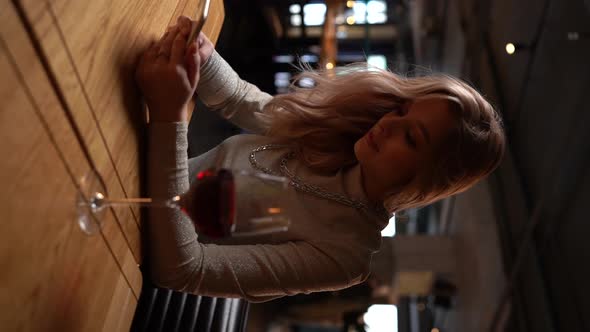 Vertical Shot of Charming Blonde Young Woman Sitting at Restaurant with Red Wine and Using Typing