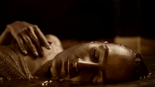 Closeup of a Mystical Young Woman Lying in a Bathtub with Water with Golden Skin Moving in a Ritual