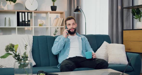 Guy Which Sitting on the Couch at Home and Talking on Smartphone Simultaneously Drinking Coffee