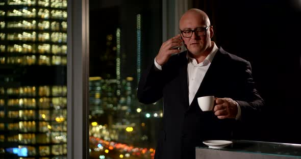 Bald Smiling Businessman in a Black Suit and Glasses Talking on the Phone, He Is in a Beautiful