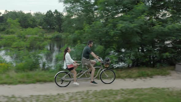 A young couple rides a tandem two-seater bike along the riverbank