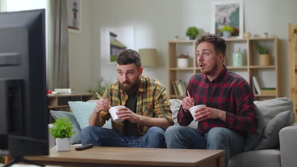 Two Young Cute Men Caucasian Eating a Chinese Noodle Sitting in a Living Room on a Couch and Watch
