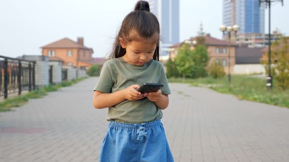 Little Asian Girl Types Message on Modern Mobile Phone Standing Outdoor