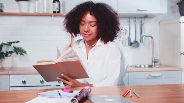 Young African American Business Woman Reads Book Tsits at Kitchen Table