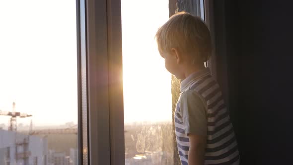 Small Blond Baby Kid Stands At Window Looks At The Street Against Sunset
