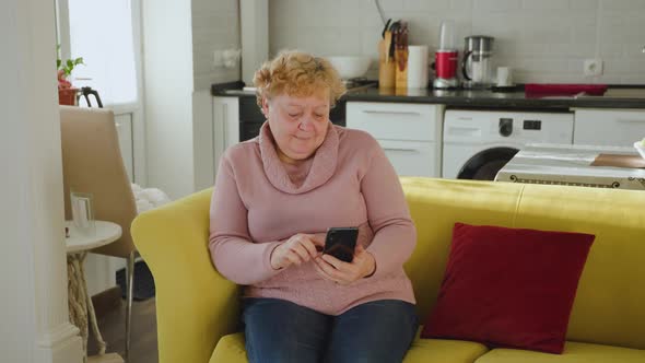 Senior Lady Using Smartphone Surfing the Net Sit on Couch at Home in the Living Room