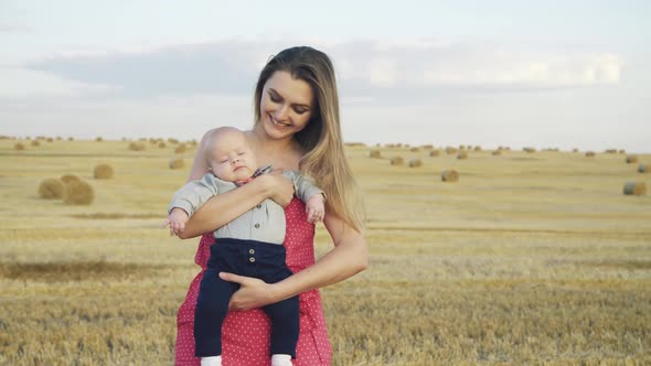 Lovely Mother Kisses and Caresses Baby Boy on Hands in Summer Wheat Hay Field