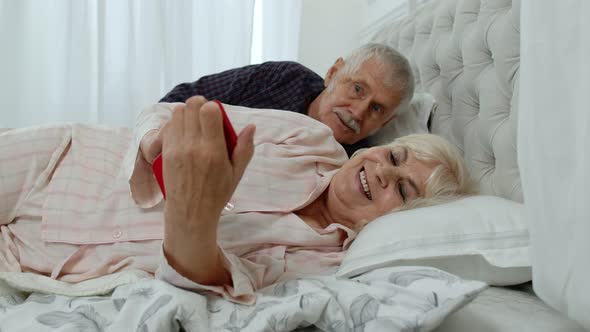 Senior Grandparents Couple Lying in Bed. Woman Getting Nervous About Man Spying Into Mobile Phone