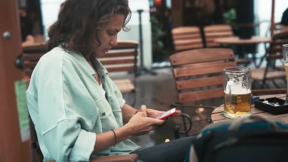 Serious Young Woman Texting with a Phone While Sitting on a Terrace of a Cafe