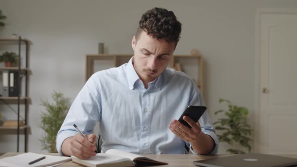 Serious Young Man in Shirt Sit at Modern Homeoffice Workplace Holding Cellphone Writing Notes in