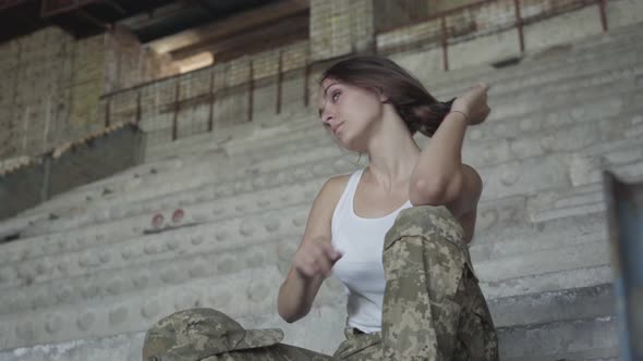 Strong Young Woman in Military Uniform Sitting on the Cold Concrete Stairs in the Abandoned Building