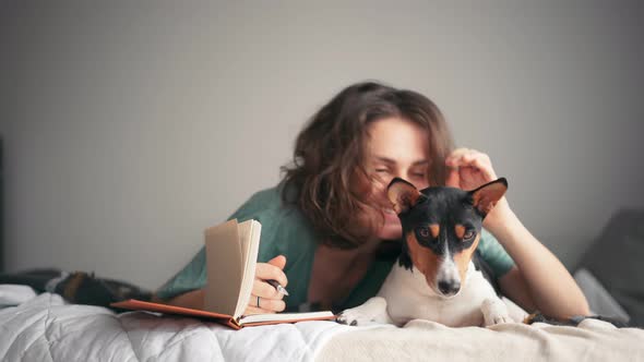 A Young Woman Is Writing a Diary While Lying in Bed with Her Basenji Dog