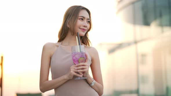 Asian woman drinking cocktail while waiting for friends at skyscraper rooftop restaurant at sunset.