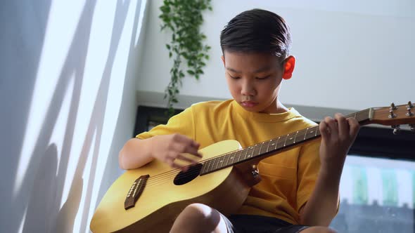 Preteen Asian boy playing acoustic guitar dressed sitting on the cozy sofa at the home living room a