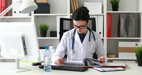 Doctor in Glasses Sitting at Table and Scrolling Folder with Documents
