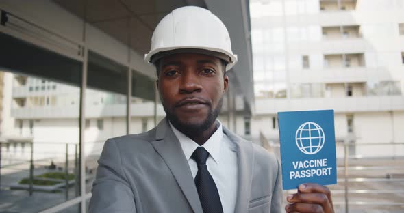 Afro Male Engineer in Hardhat Looking at Camera and Talking Holding Vaccination Certificate