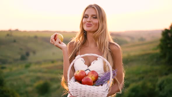 Beautiful sexy blonde girl in white dress posing in a field at sunset with a basket of fruit