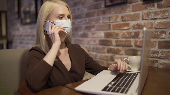 Young Woman Manager Answers a Phone Call and Types Text on a Laptop Keyboard in a Cafe