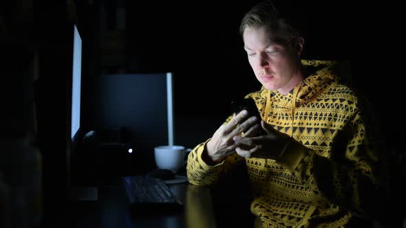 Young Man Using Phone and Drinking Coffee While Working Overtime at Home in the Dark