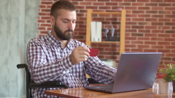 Disabled Person is Sitting Wheelchair Front of Laptop and Typing Something with Bank Card in His