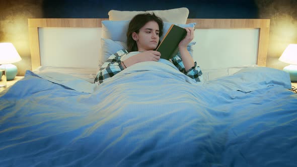 Brunette Girl in the Evening Lying in Her Bed Reading a Book Front View