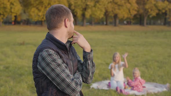 Thoughtful Caucasian Man Waving To His Daughters on the Background and Looking at the Camera