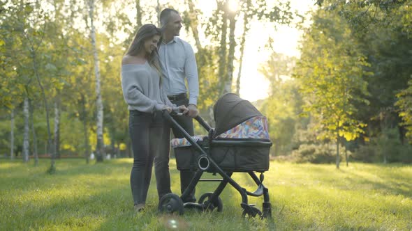 Wide Shot of Happy Smiling Husband and Wife Standing with Baby Stroller in Sunny Park and Talking