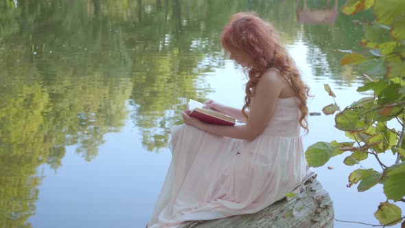 Redhead Attractive Caucasian Girl Reading the Book in Dark Red Cover on the Bank of the Lake in the
